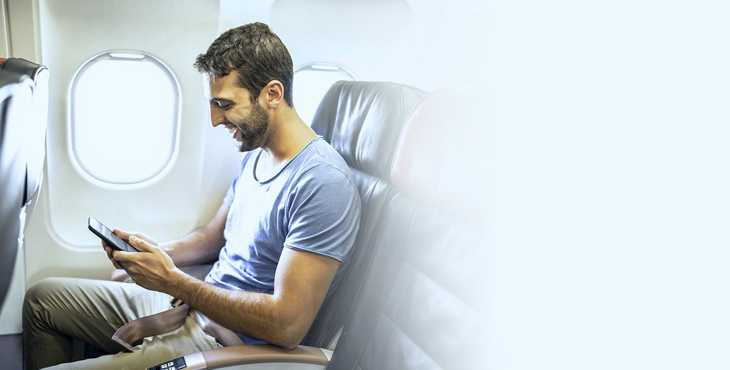 Man with brown hair and a beard sitting next to a window on a plane looking at his smartphone
