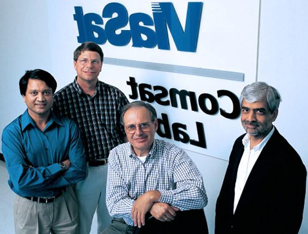 在Viasat通信卫星实验室标志前，四名Viasat员工对着摄像机微笑