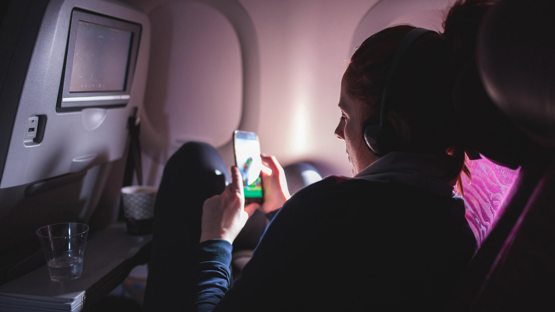 一个戴着耳机的人蜷缩在飞机座位上，看着手机屏幕上的东西. 
